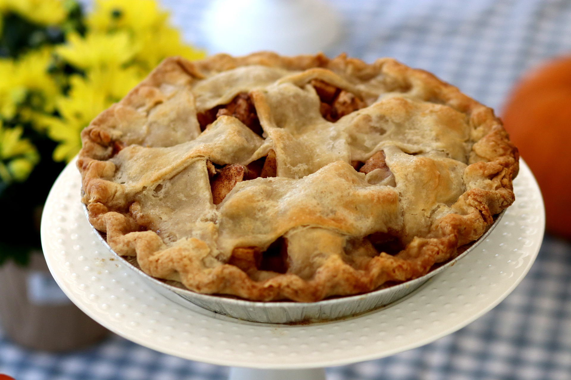 Lattice-crust apple pie on a white pedestal stand with mums and a pumpkin