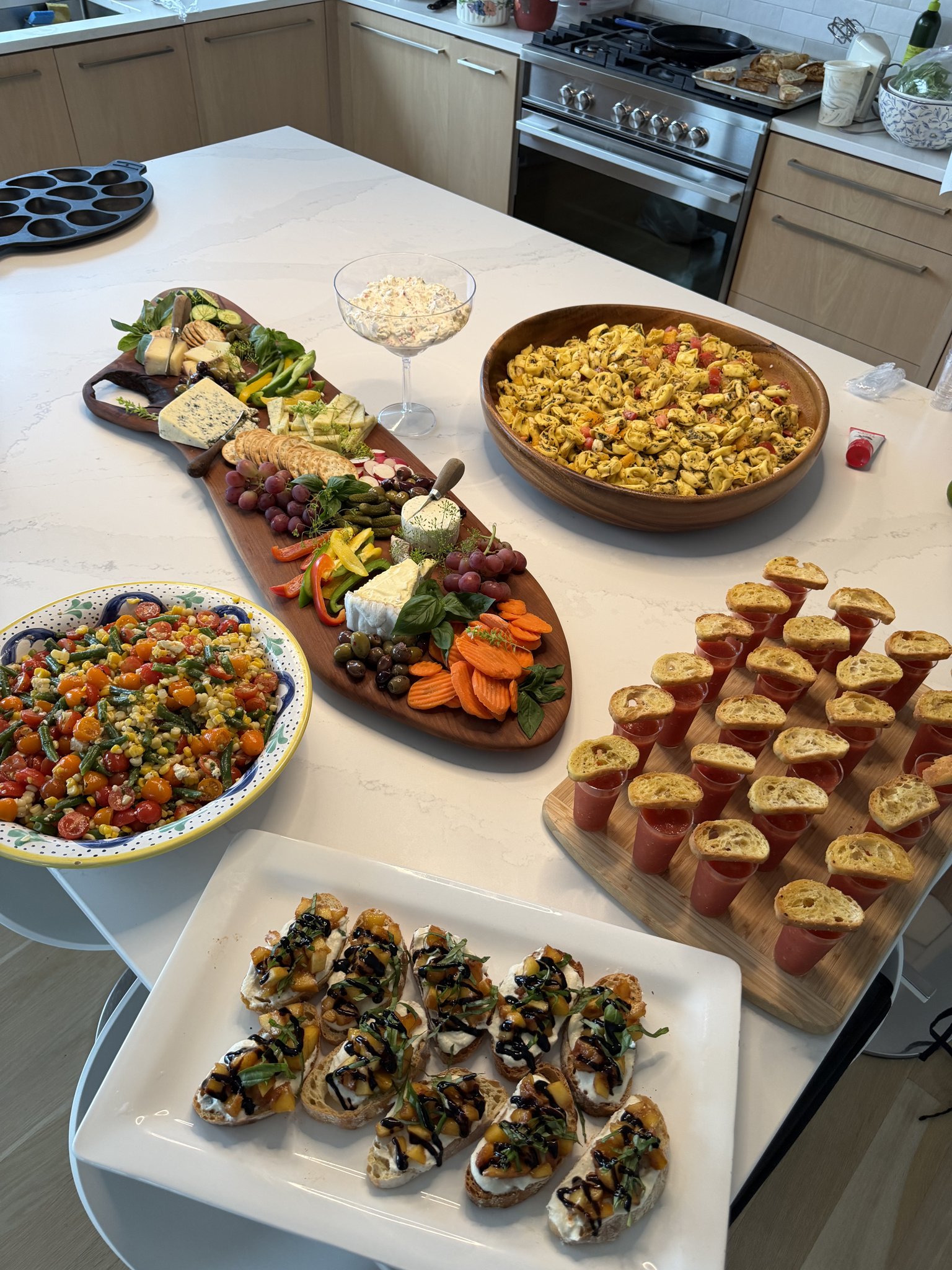 A catered spread on a marble island — grazing board, summer salad, bruschetta and gazpacho shooters