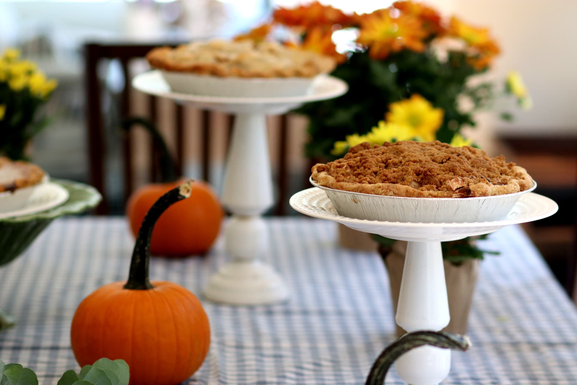 Apple crumble pie on a white pedestal stand with pumpkins and mums on a blue gingham tablecloth